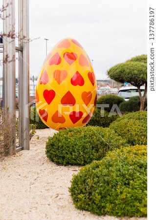 A big yellow Easter egg covered in red heart shapes is placed in a green garden near small bushes. It is daytime and shoppers can be seen in the background of the scene A big yellow Easter egg covered in red heart shapes is placed in a green garden near small bushes. It is daytime and shoppers can be seen in the background of the scene 137786197