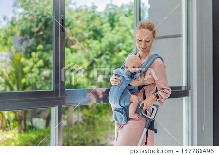 Woman vacuuming a room while carrying her baby in an ergonomic carrier. Modern motherhood, multitasking parent, home chores with infant, balance of childcare and housework concept. Woman vacuuming a room while carrying her baby in an ergonomic carrier. Modern motherhood, multitasking parent, home chores with infant, balance of childcare and housework concept. 137786496