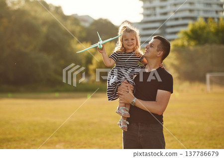 Man is holding girl high, she is with toy plane. Father with his little daughter are on the field 137786679