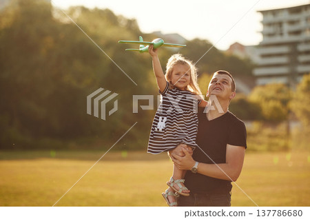 Man is holding girl high, she is with toy plane. Father with his little daughter are on the field 137786680