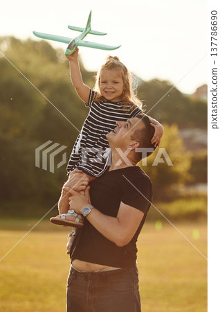 Toy plane is high up in hand of girl. Father with his little daughter are on the field Toy plane is high up in hand of girl. Father with his little daughter are on the field 137786690