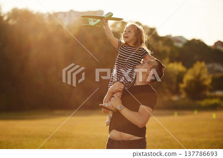 Toy plane is high up in hand of girl. Father with his little daughter are on the field Toy plane is high up in hand of girl. Father with his little daughter are on the field 137786693