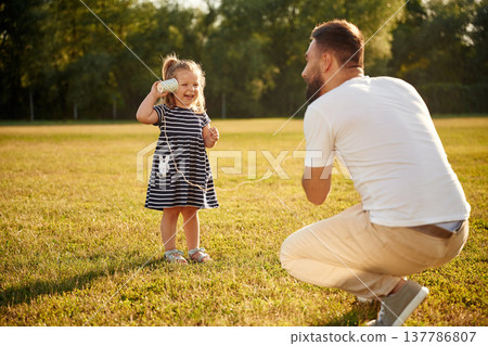 Speaking by using tin can telephone. Father with his little daughter are on the field Speaking by using tin can telephone. Father with his little daughter are on the field 137786807