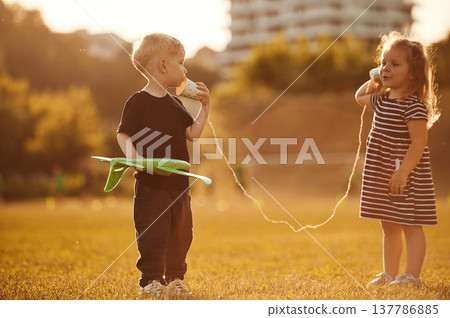 Conversation by using tin can telephone. Two kids are having fun on the summer field Conversation by using tin can telephone. Two kids are having fun on the summer field 137786885