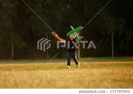 Running with two toy planes in hands. Boy is having fun on the field at daytime Running with two toy planes in hands. Boy is having fun on the field at daytime 137787043