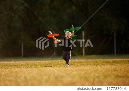 Running with two toy planes in hands. Boy is having fun on the field at daytime Running with two toy planes in hands. Boy is having fun on the field at daytime 137787044