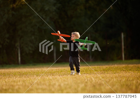 Running with two toy planes in hands. Boy is having fun on the field at daytime Running with two toy planes in hands. Boy is having fun on the field at daytime 137787045