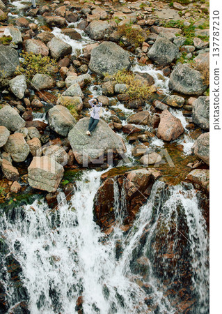 A photographer girl sits on a rock at the edge of the Battery waterfall in Teriberka, Arctic landscape with a view of a flowing waterfall in the northern fjord, Arctic, Russia, Murmansk region A photographer girl sits on a rock at the edge of the Battery waterfall in Teriberka, Arctic landscape with a view of a flowing waterfall in the northern fjord, Arctic, Russia, Murmansk region 137787210