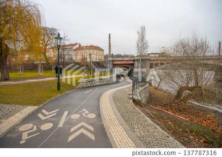 A bicycle path curves along the river in Prague. The scene shows a route marked for cyclists. Nearby, a bridge crosses the water. The sky appears gray, suggesting a cool day. 137787316