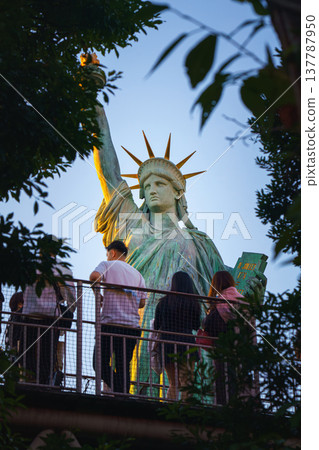 Tokyo, Japan - Oct 13 2024, vertical view of Tokyo Statue of Liberty on embankment on Odaiba Island, through foliage of trees and people on foreground, at daytime with blue sky, Tokyo, Japan Tokyo, Japan - Oct 13 2024, vertical view of Tokyo Statue of Liberty on embankment on Odaiba Island, through foliage of trees and people on foreground, at daytime with blue sky, Tokyo, Japan 137787950