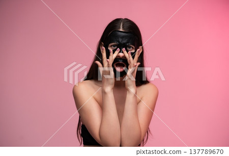 Young woman surprised while removing a charcoal face mask, revealing clear, glowing skin during a self-care beauty routine on a pink studio background 137789670