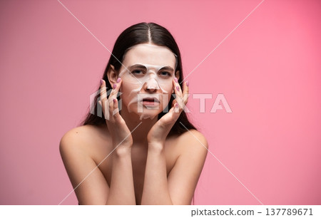 Woman applying a hydrating sheet mask to her face, promoting skincare and self-care routines for beauty, wellness, and healthy glowing skin against a pink background 137789671