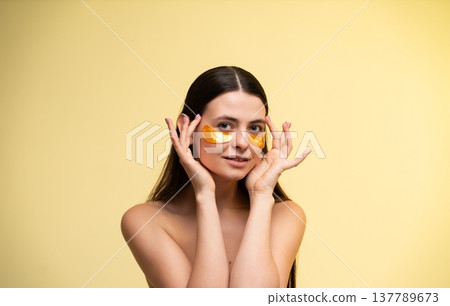 Young woman applying golden eye patches for under-eye skincare treatment, focusing on beauty, hydration, and self-care routine with glowing skin on yellow background Young woman applying golden eye patches for under-eye skincare treatment, focusing on beauty, hydration, and self-care routine with glowing skin on yellow background 137789673