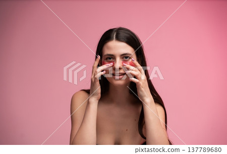 Young woman applying red hydrogel eye patches for skincare treatment and beauty routine, smiling at the camera on a vibrant pink studio background Young woman applying red hydrogel eye patches for skincare treatment and beauty routine, smiling at the camera on a vibrant pink studio background 137789680