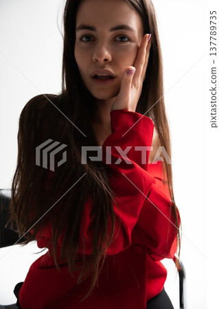 Young woman with long dark hair covering part of her face with a hand, looking vulnerable, dressed in a stylish red blazer, posing for a studio portrait on a white background 137789735