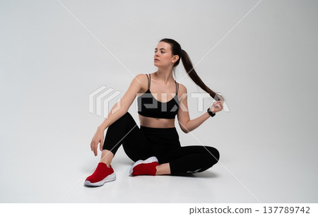 Fitness woman sitting on white background, wearing black sportswear and red sneakers, pulling her long ponytail, demonstrating a healthy and active lifestyle 137789742