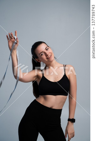 Woman standing against a gray background, smiling while holding up a measuring tape, representing health, diet, weight loss, fitness, and successful self-improvement goals Woman standing against a gray background, smiling while holding up a measuring tape, representing health, diet, weight loss, fitness, and successful self-improvement goals 137789743
