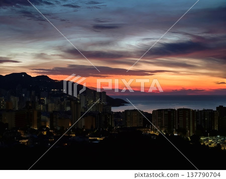 Gentle Evening Scene Of Waterfront With Shimmering Lights And Highrise Silhouettes Across Calm Waters 137790704