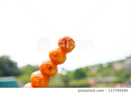 A close-up shot of a mitarashi dango skewer against a backdrop of blue sky and nature. This traditional Japanese confectionary shines with its sweet and savory sauce. A close-up shot of a mitarashi dango skewer against a backdrop of blue sky and nature. This traditional Japanese confectionary shines with its sweet and savory sauce. 137794490
