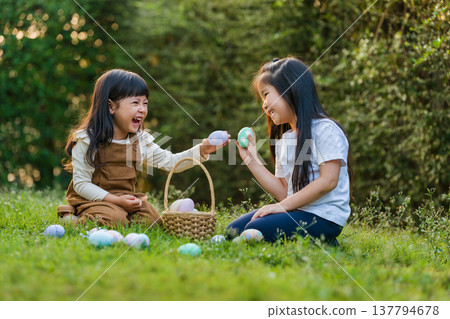 two happy child girl playing together and hunting for Easter eggs into basket on green grass in garden two happy child girl playing together and hunting for Easter eggs into basket on green grass in garden 137794678