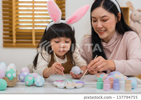 mother and preschool child girl wearing bunny ears painting and decorating egg together at home on easter day. happy family holiday activity concept mother and preschool child girl wearing bunny ears painting and decorating egg together at home on easter day. happy family holiday activity concept 137794755