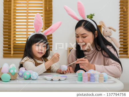mother and preschool child girl wearing bunny ears painting and decorating egg together at home on easter day. happy family holiday activity concept mother and preschool child girl wearing bunny ears painting and decorating egg together at home on easter day. happy family holiday activity concept 137794756