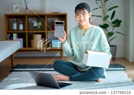 A smiling woman holding a calculator and a box; an image of selling items on a flea market app, running an online shop, or a side hustle. A smiling woman holding a calculator and a box; an image of selling items on a flea market app, running an online shop, or a side hustle. 137798510