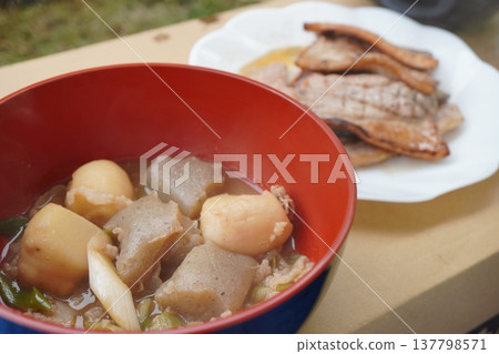 A scene of a dining table with a warm stew containing taro, konjac, and vegetables, alongside plates of grilled meat. A scene of a dining table with a warm stew containing taro, konjac, and vegetables, alongside plates of grilled meat. 137798571