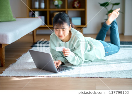 A young woman is relaxing in her living room and online shopping on her laptop. A young woman is relaxing in her living room and online shopping on her laptop. 137799103