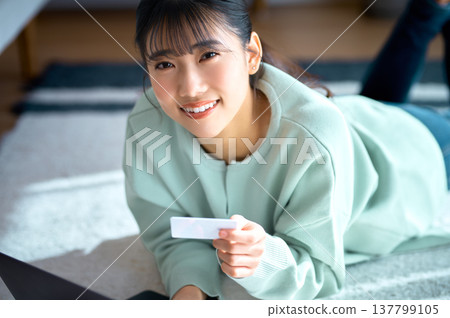一位年輕女子麵帶微笑,手持信用卡,一邊進行網上購物一邊看向鏡頭。 一位年輕女子麵帶微笑,手持信用卡,一邊進行網上購物一邊看向鏡頭。 137799105
