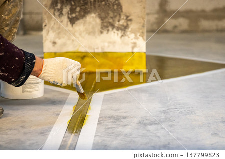 Detailed shot of a hand in a glove applying yellow paint to a floor joint using a brush and masking tape for precision. Detailed shot of a hand in a glove applying yellow paint to a floor joint using a brush and masking tape for precision. 137799823