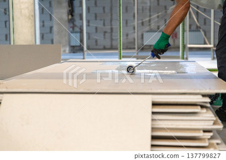 Close-up of a worker's hand in a green glove using a paint roller to apply white primer or adhesive to stacked building boards. Close-up of a worker's hand in a green glove using a paint roller to apply white primer or adhesive to stacked building boards. 137799827