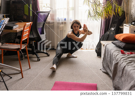 A woman performs a deep side lunge stretch in her room, focusing on flexibility and leg strength. 137799831