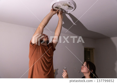 A man holds two light bulbs while installing them into a ceiling light fixture during a home maintenance task. A man holds two light bulbs while installing them into a ceiling light fixture during a home maintenance task. 137799852