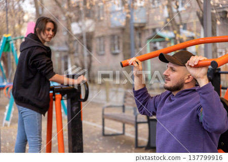 Man performing strength exercises on a public gym machine while talking to a female friend outdoors. 137799916