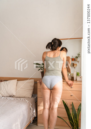 Woman standing in bedroom, looking at her body reflection in the dresser mirror, practicing self-care and body positivity. Adult woman admiring body reflecting in mirror at home 137800244