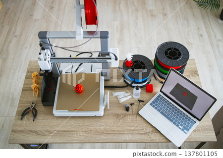 A 3D printer is set up on a wooden desk with spools of filament, tools, and a laptop displaying a 3D model A 3D printer is set up on a wooden desk with spools of filament, tools, and a laptop displaying a 3D model 137801105