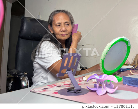Senior woman sitting at desk and adjusting her hair in front of mirror, elderly lady doing self-care routine in her room Senior woman sitting at desk and adjusting her hair in front of mirror, elderly lady doing self-care routine in her room 137801113