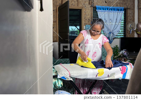Senior woman ironing clothes on board in authentic home interior, elderly lady doing domestic chores, household routine portrait Senior woman ironing clothes on board in authentic home interior, elderly lady doing domestic chores, household routine portrait 137801114