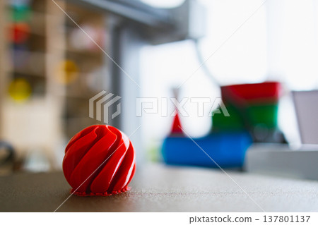 A bright red, intricately designed sphere sits on the print bed of a 3D printer, with other colorful objects blurred in the background A bright red, intricately designed sphere sits on the print bed of a 3D printer, with other colorful objects blurred in the background 137801137