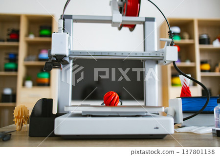 A modern 3D printer is actively creating a red, spiral-shaped object on its build plate, with spools of filament visible in the background A modern 3D printer is actively creating a red, spiral-shaped object on its build plate, with spools of filament visible in the background 137801138