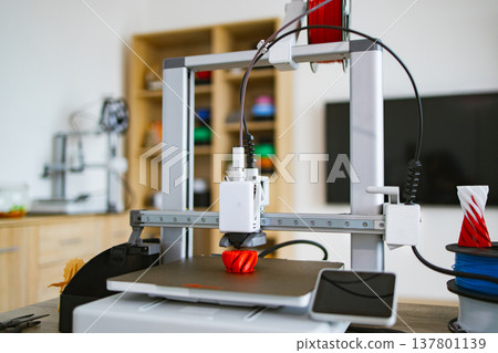 A modern 3D printer is actively creating a red object on its build plate with filament spools nearby A modern 3D printer is actively creating a red object on its build plate with filament spools nearby 137801139