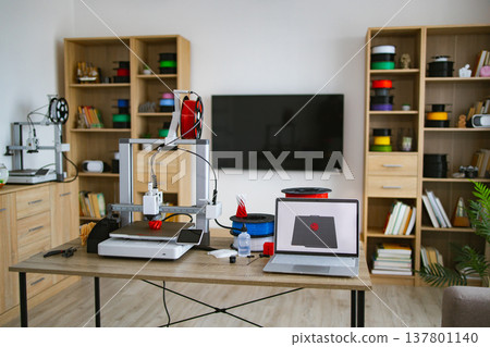 A modern 3D printing workstation is set up on a desk with multiple printers, filament spools, and a laptop displaying a design A modern 3D printing workstation is set up on a desk with multiple printers, filament spools, and a laptop displaying a design 137801140