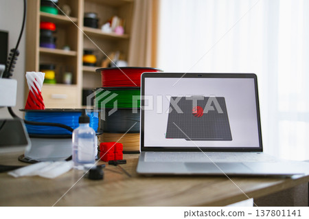 A modern laptop displays 3D modeling software next to spools of filament and a printed object, showcasing a home fabrication workspace A modern laptop displays 3D modeling software next to spools of filament and a printed object, showcasing a home fabrication workspace 137801141
