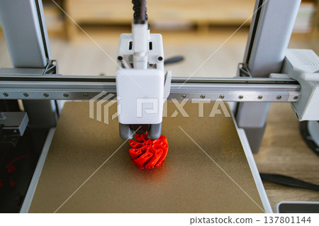 A close-up view of a 3D printer's nozzle depositing molten plastic to build a complex red object layer by layer on a gold build plate A close-up view of a 3D printer's nozzle depositing molten plastic to build a complex red object layer by layer on a gold build plate 137801144