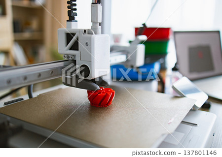 A modern 3D printer is actively creating a small, intricate red object on its build plate with a laptop and smartphone in the background 137801146