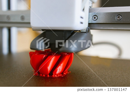 A close-up view of a 3D printer nozzle depositing molten plastic to build a red, intricate object layer by layer 137801147