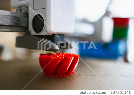 A 3D printer nozzle extrudes molten plastic to build a vibrant red object layer by layer on a textured build plate A 3D printer nozzle extrudes molten plastic to build a vibrant red object layer by layer on a textured build plate 137801148
