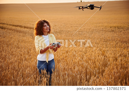 Modern black drone is up. Beautiful woman is on the wheat agricultural field Modern black drone is up. Beautiful woman is on the wheat agricultural field 137802371