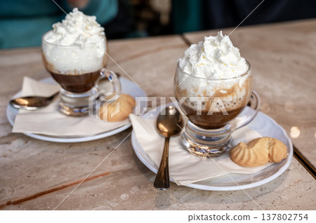 Two clear glasses filled with layered coffee and generously topped with fluffy whipped cream, served with small cookies on saucers and spoons on a marble table 137802754